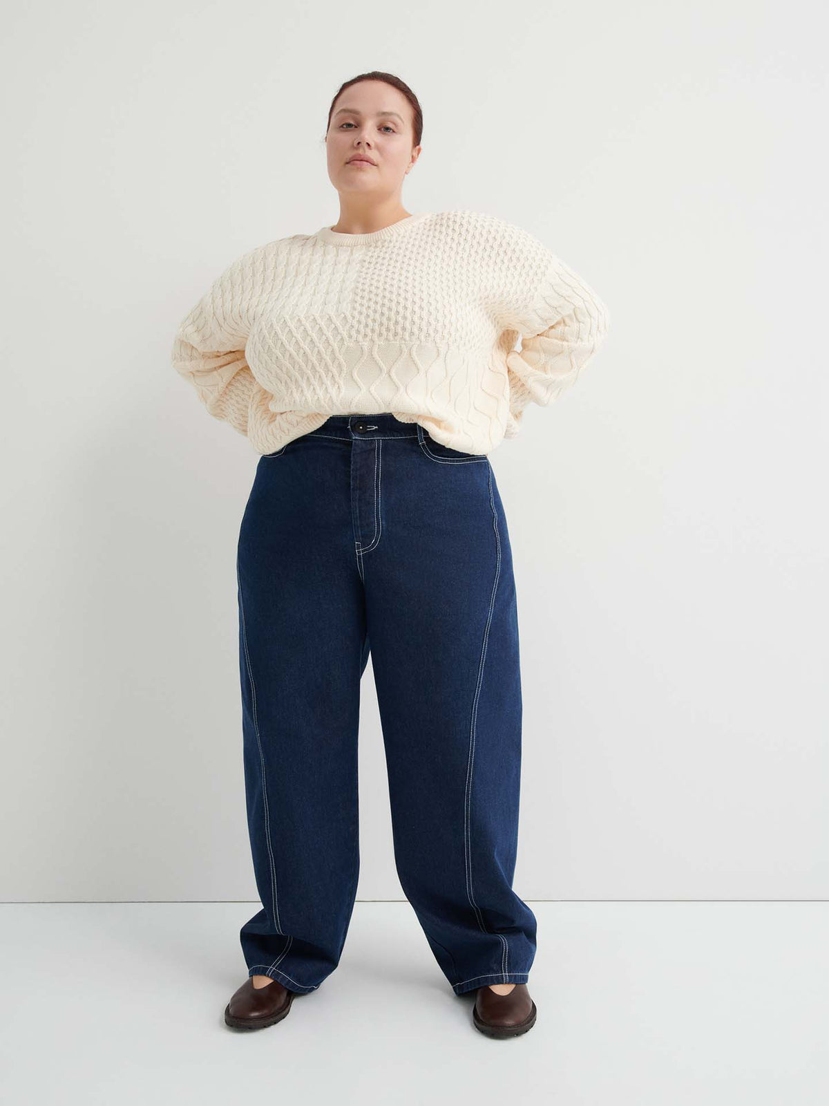 A person stands against a plain light-colored wall, wearing a cream textured sweater, indigo denim Twist Jeans – Indigo Denim in a relaxed high-waisted fit, and brown shoes.