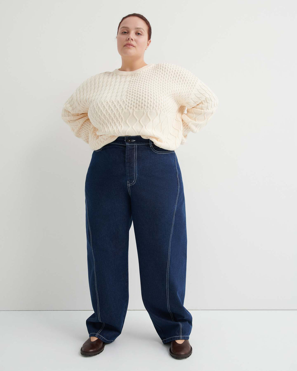 A person stands against a plain light-colored wall, wearing a cream textured sweater, indigo denim Twist Jeans – Indigo Denim in a relaxed high-waisted fit, and brown shoes.