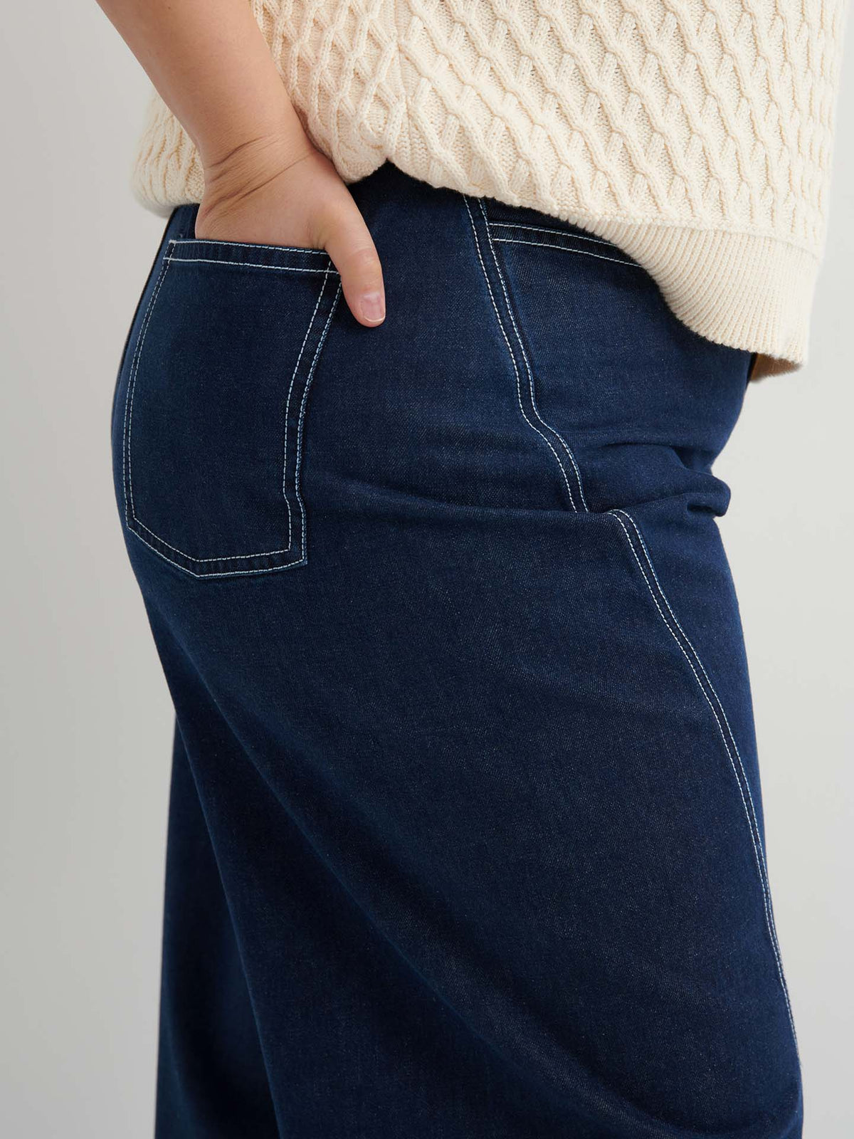 Close-up of a person’s hand in the back pocket of Twist Jeans – Indigo Denim, paired with a cream textured sweater.