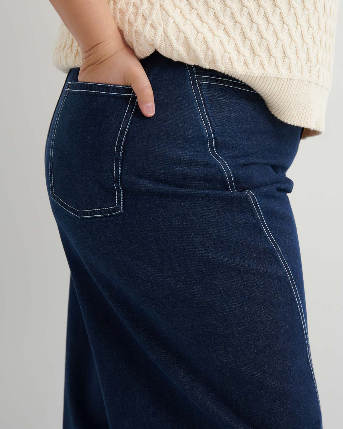 Close-up of a person’s hand in the back pocket of Twist Jeans – Indigo Denim, paired with a cream textured sweater.