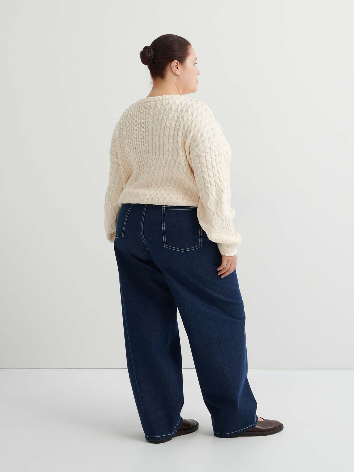 A person stands facing away, wearing a cream cable knit sweater, Twist Jeans – Indigo Denim with visible stitching and relaxed fit, and dark shoes, against a plain white background.