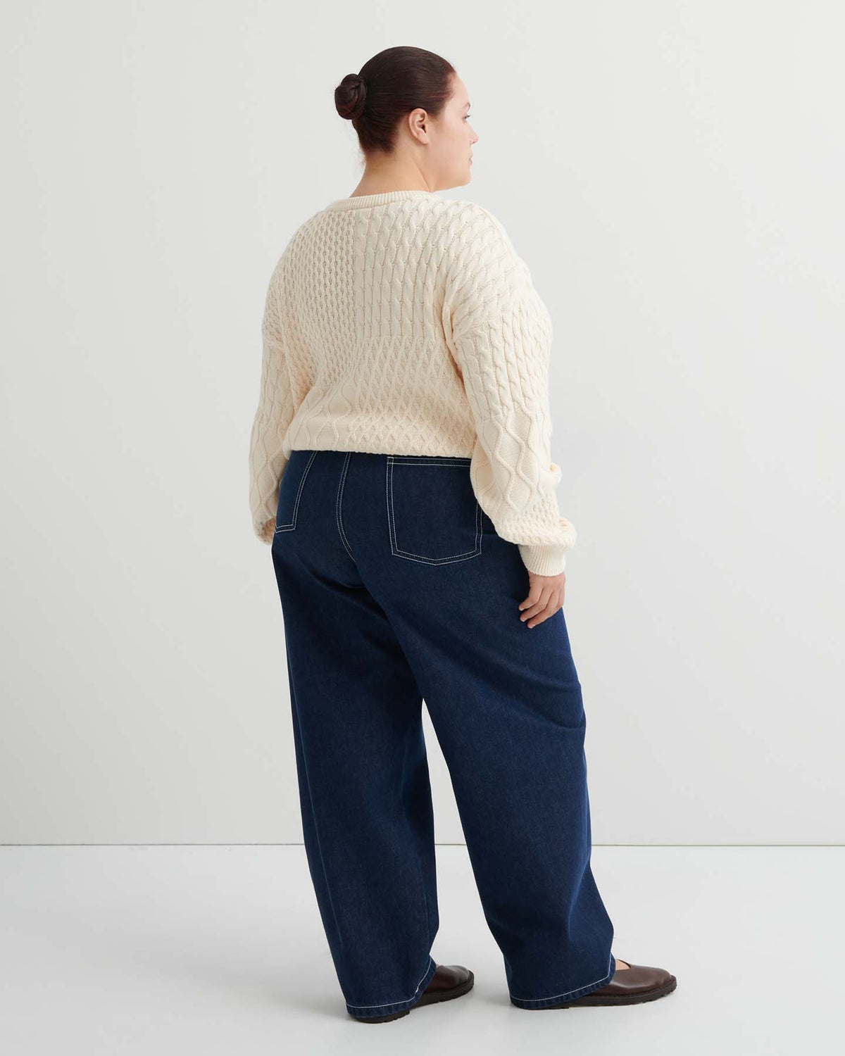A person stands facing away, wearing a cream cable knit sweater, Twist Jeans – Indigo Denim with visible stitching and relaxed fit, and dark shoes, against a plain white background.