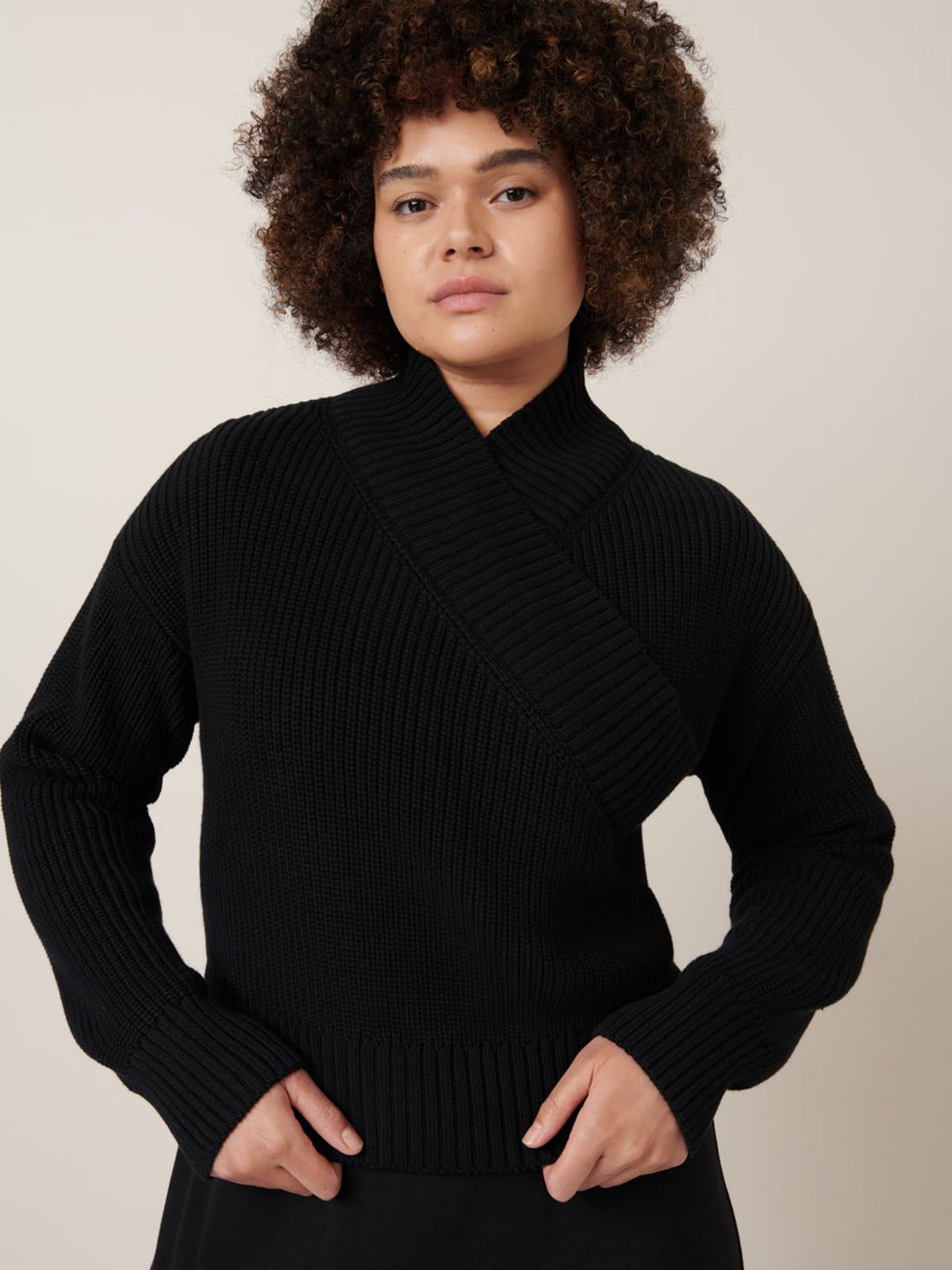 A person with curly hair wears the Kowtow Unity Cardigan in black, featuring ribbed knitwear and wrap ties, and looks at the camera with a neutral expression against a plain background.