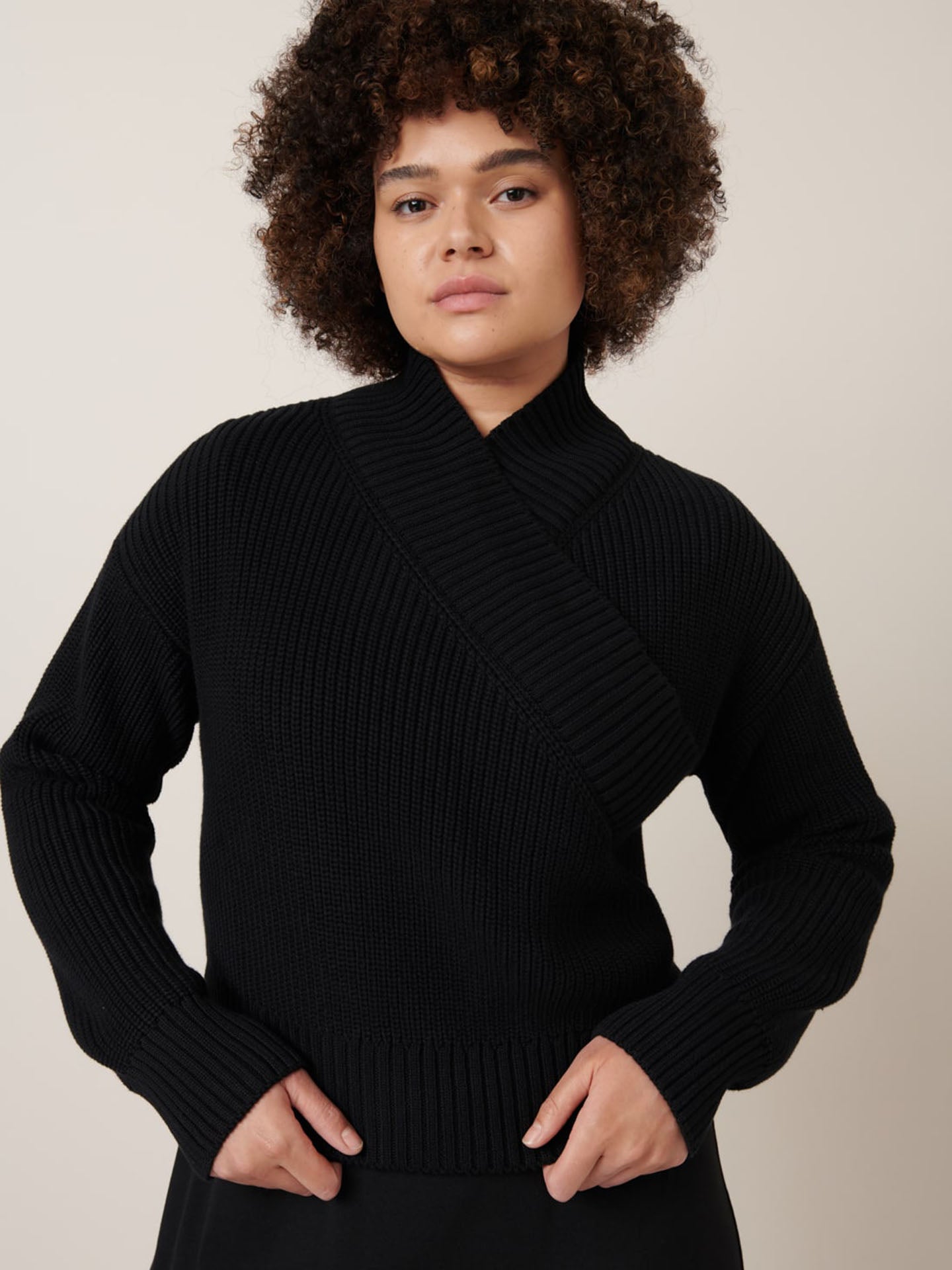 A person with curly hair wears the Kowtow Unity Cardigan in black, featuring ribbed knitwear and wrap ties, and looks at the camera with a neutral expression against a plain background.