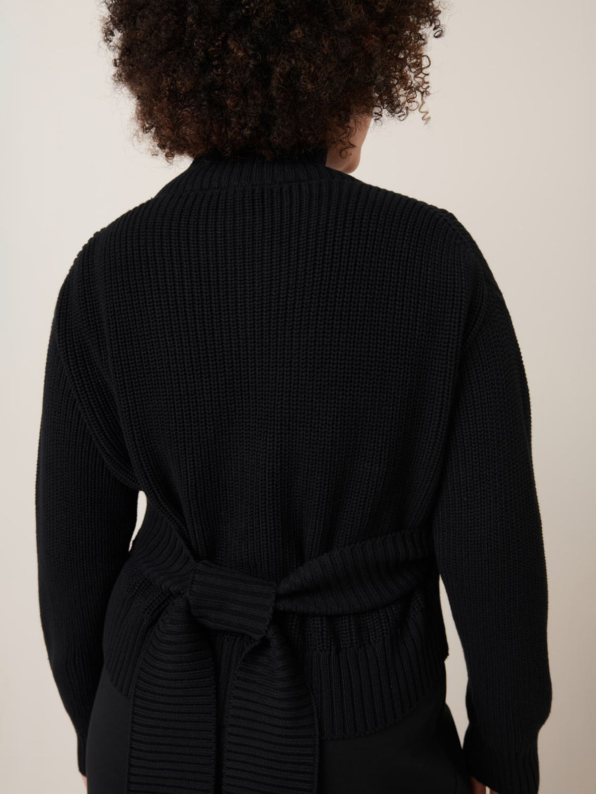 A person seen from the back wearing the Kowtow Unity Cardigan in black, a long-sleeve knitwear piece featuring wrap ties and a belted waist detail.