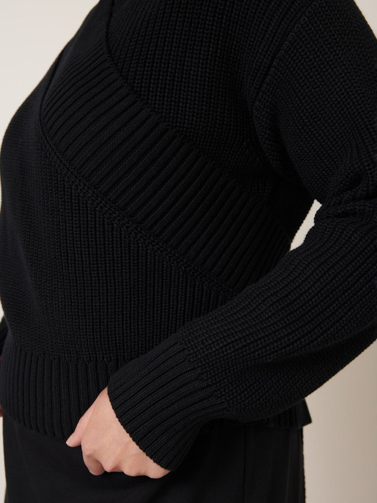 Model wears the Unity Cardigan – Black by Kowtow, featuring ribbed and diagonal patterns in textured knit. Shown from the side with one hand near the hip. Check our size guide for your perfect fit.