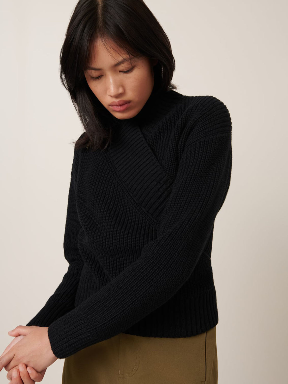 A person with straight black hair, wearing the Kowtow Unity Cardigan in black and khaki pants, looks downward with hands clasped.