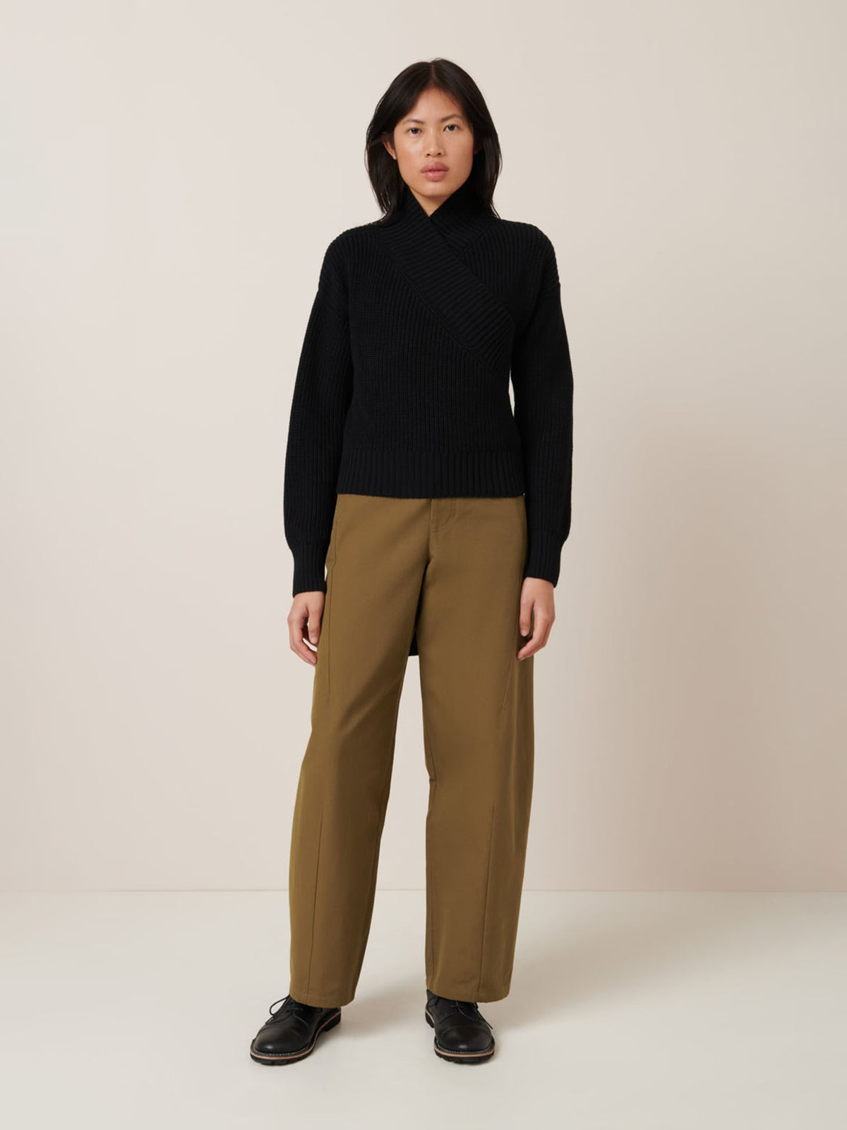 A person stands facing forward against a plain light background, wearing the Kowtow Unity Cardigan in black, paired with olive-green pants and black shoes.