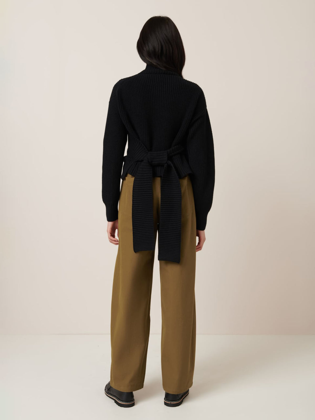 A person stands facing away against a plain light background, wearing Kowtow&#39;s Unity Cardigan in black with wrap ties at the back, paired with wide-leg khaki pants and black shoes.