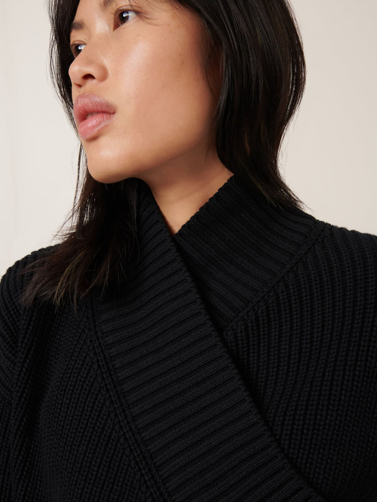 A person with long dark hair wears the Kowtow Unity Cardigan in black and looks off to the side against a neutral background.