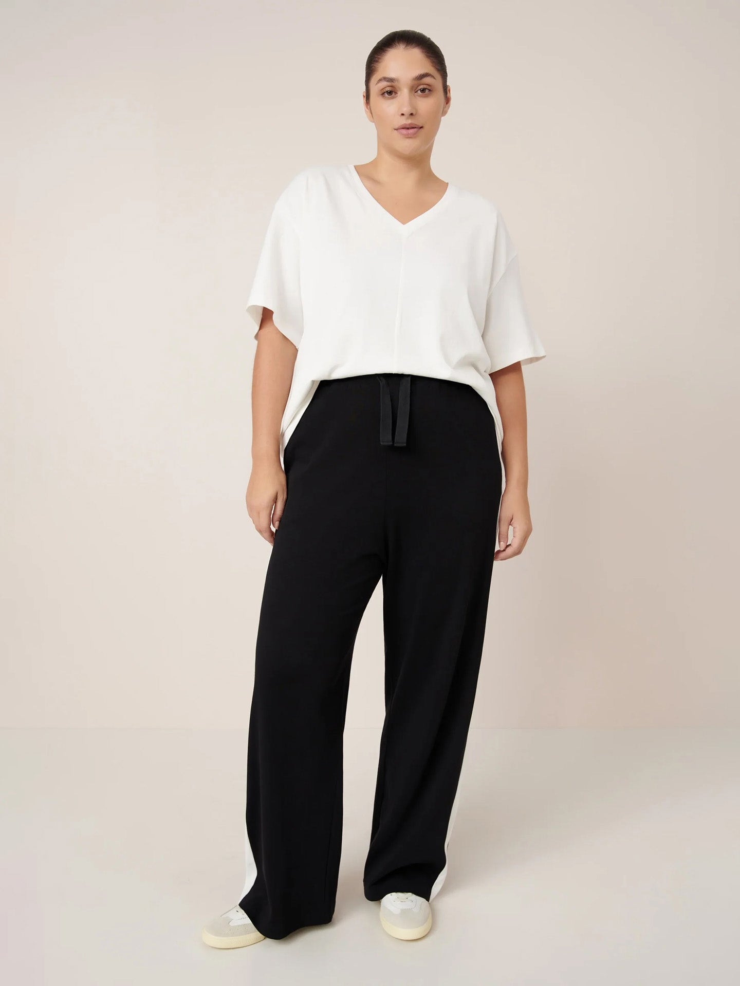 A person stands facing forward, wearing the Kowtow Unity Tee in Off White, black wide-leg pants with a tie waist, and white sneakers, against a plain beige background.