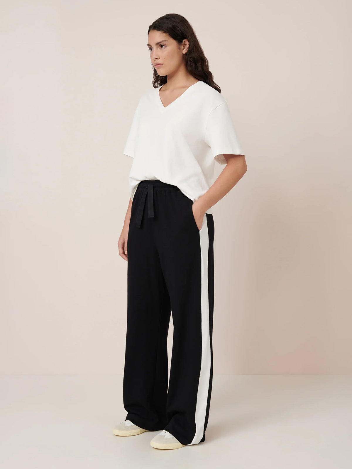 A person wears the Kowtow Unity Tee – Off White, paired with wide-leg black pants featuring white side stripes and white sneakers, against a plain background. Garment measurements are available for accurate size selection.