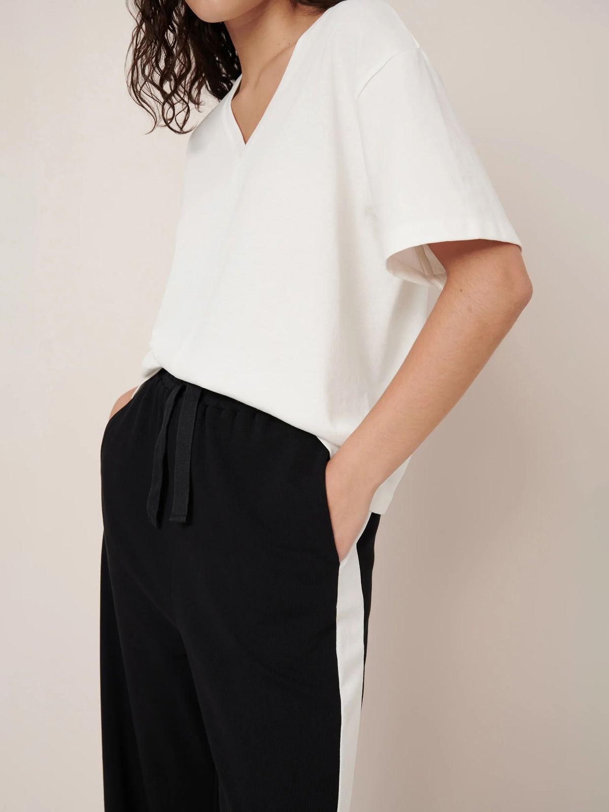 Someone stands with hands in pockets against a plain background, wearing the Kowtow Unity Tee in Off White and black relaxed-fit pants with white side stripes.