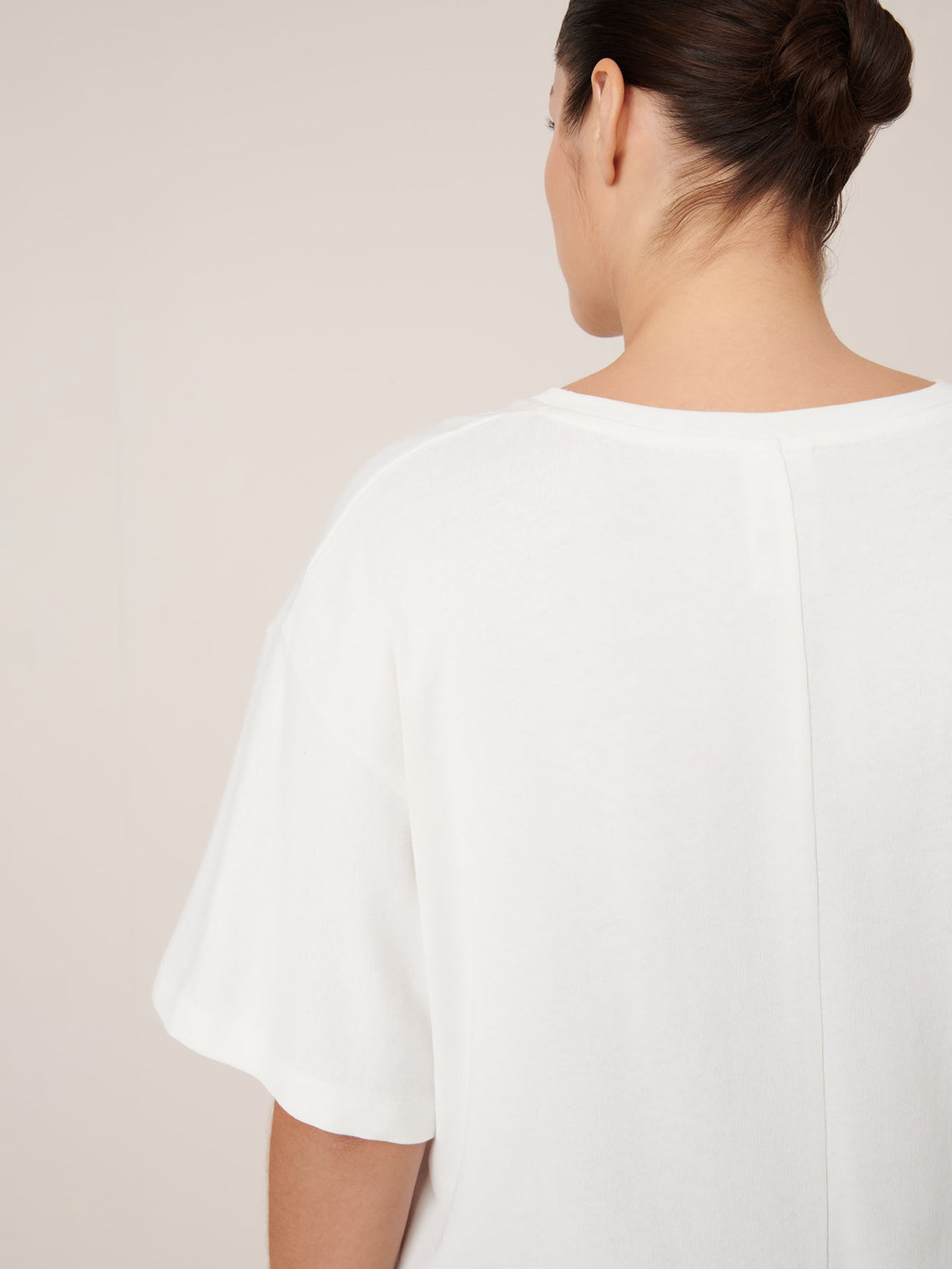 A person with dark hair in a bun is shown from the back wearing Kowtow’s Unity Tee in Off White, a relaxed fit short-sleeve top, against a plain light background.