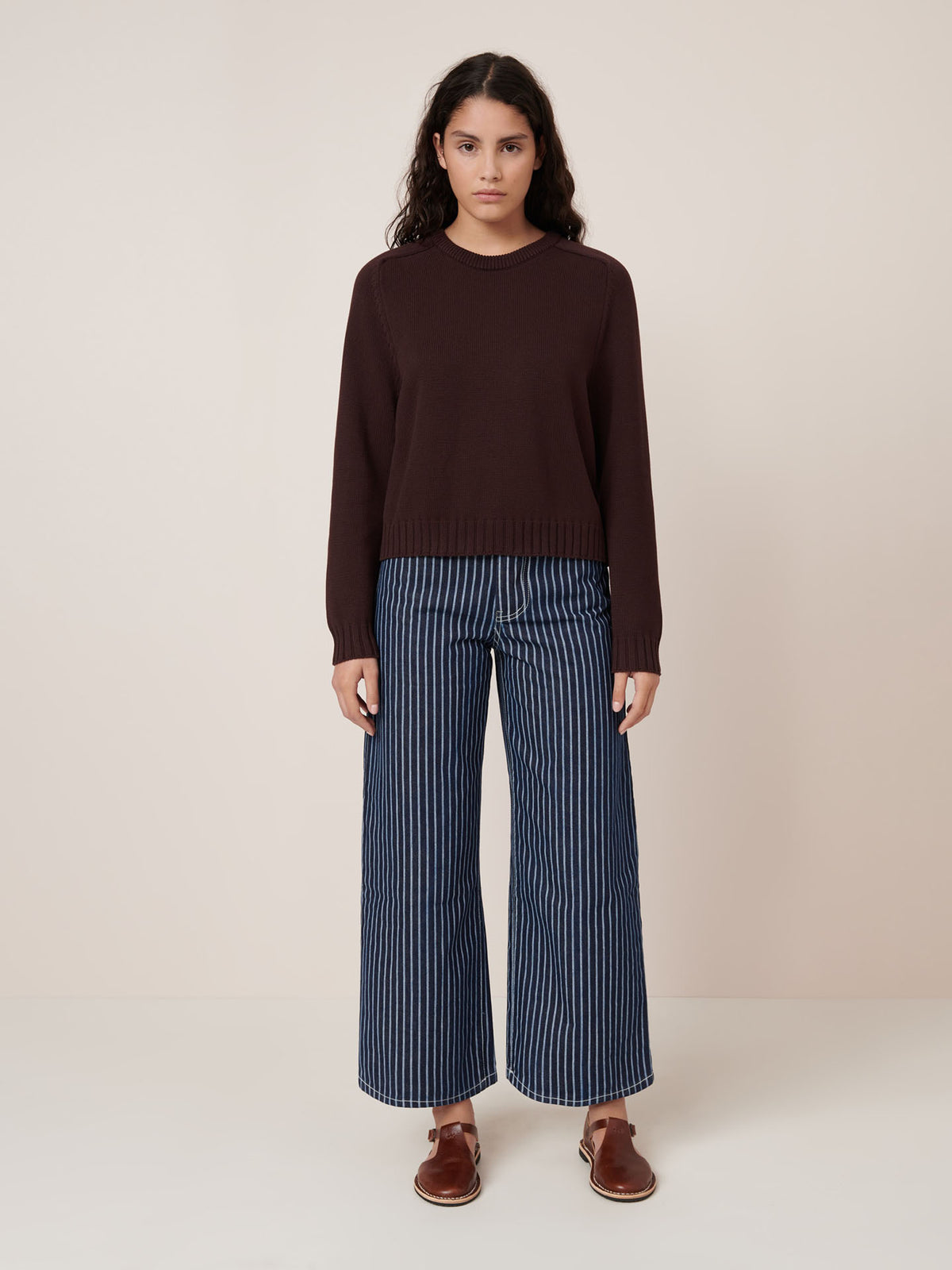A person stands against a plain background wearing the Kowtow Valley Jumper in Mahogany, blue and white vertical striped wide-leg pants with a relaxed fit, and brown sandals.