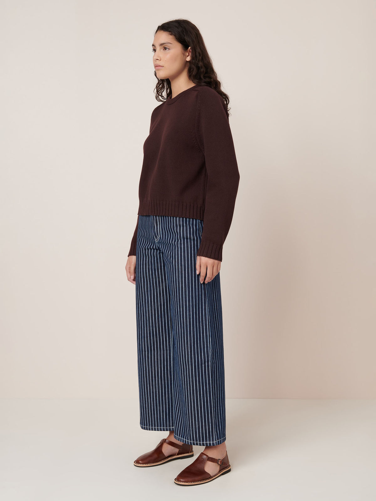 A person stands against a plain background wearing the Kowtow Valley Jumper in mahogany, blue and white vertically striped relaxed fit wide-leg pants, and brown flat shoes.