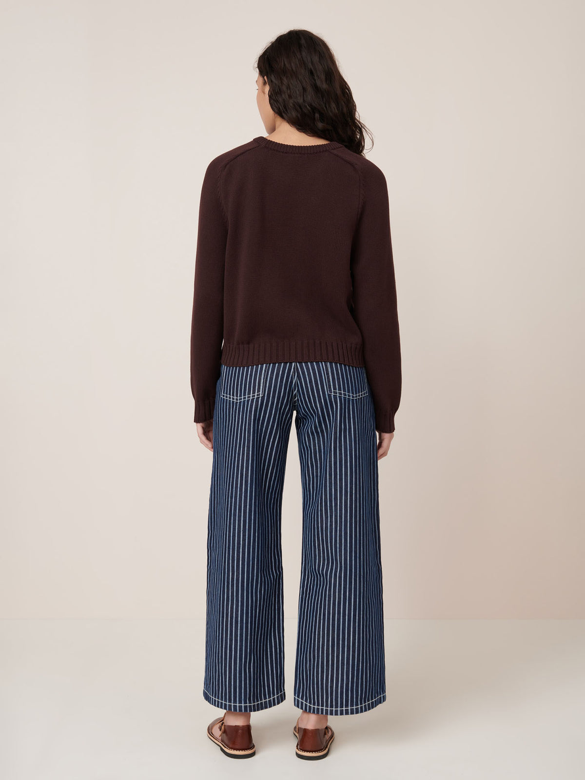 Person stands with back to camera wearing Kowtow’s Valley Jumper in Mahogany and blue and white vertical striped wide-leg pants, paired with sandals against a plain background.