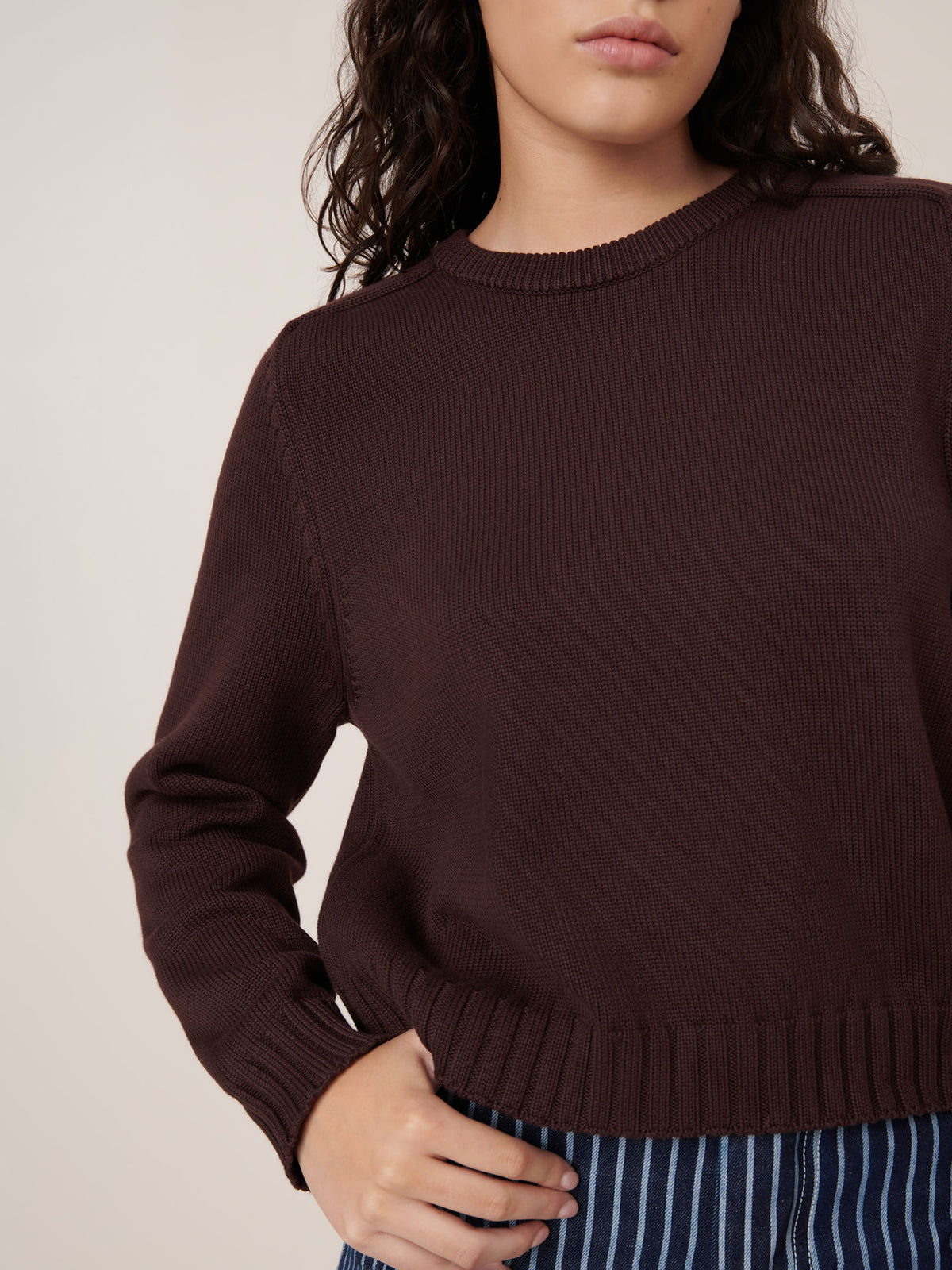 A person is shown from shoulders to hips wearing the Kowtow Valley Jumper in Mahogany, a dark brown knit sweater with a relaxed fit, paired with blue striped pants, set against a plain background.