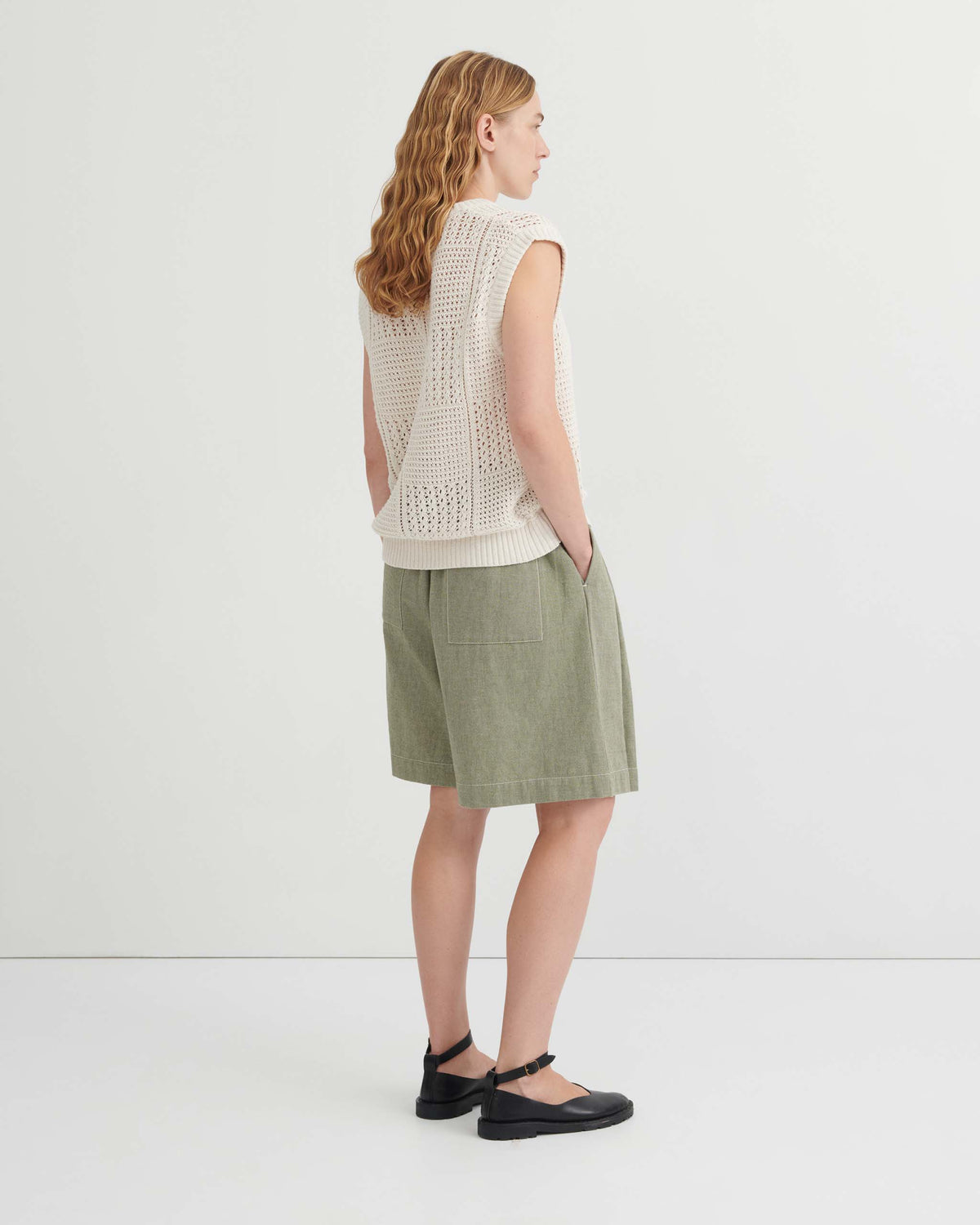 A person seen from behind wearing an ivory crochet sweater vest, olive green shorts, and black flat shoes.