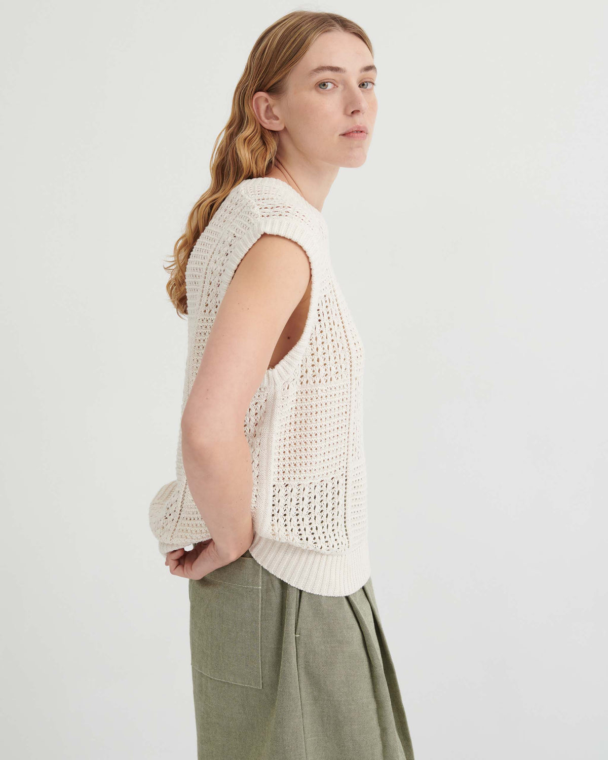 A person wearing an ivory, sleeveless, open-knit sweater vest and olive green trousers.