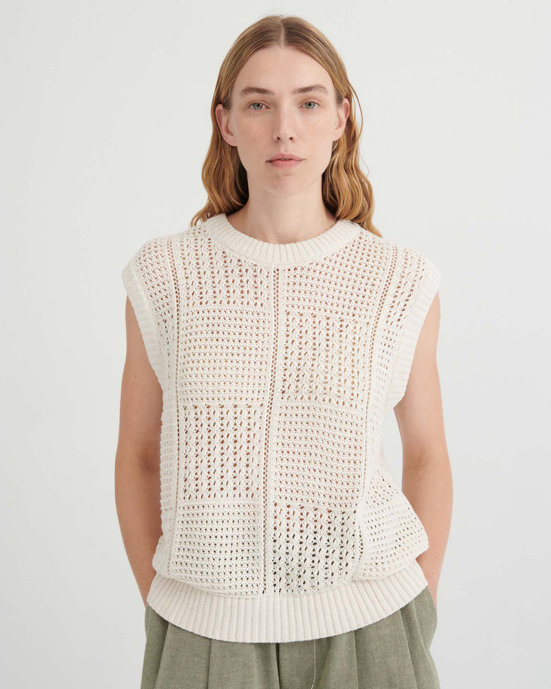 A person wearing a sleeveless, cream-colored crochet knit top with a square-patterned texture.