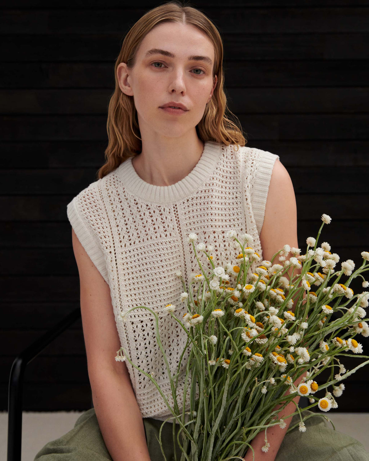 A woman wearing a cream-colored knit sleeveless top holds a bouquet of white and yellow daisies.