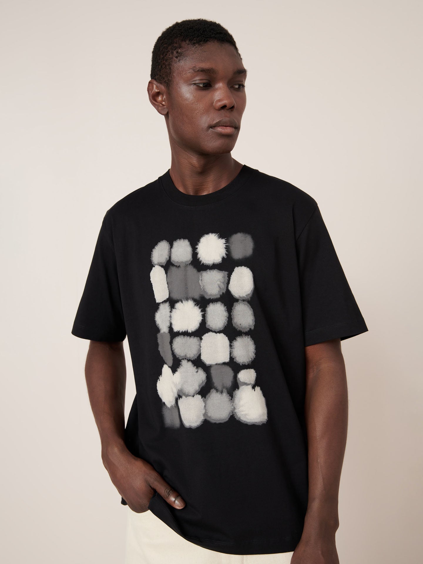 A man wears the Kowtow Watercolour Tee – Black, a standard-fit t-shirt featuring an abstract white and gray rectangular pattern, standing against a plain light background.