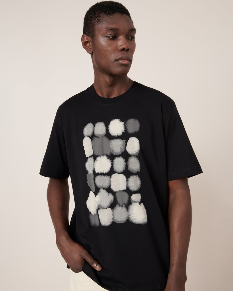 A man wears the Kowtow Watercolour Tee – Black, a standard-fit t-shirt featuring an abstract white and gray rectangular pattern, standing against a plain light background.