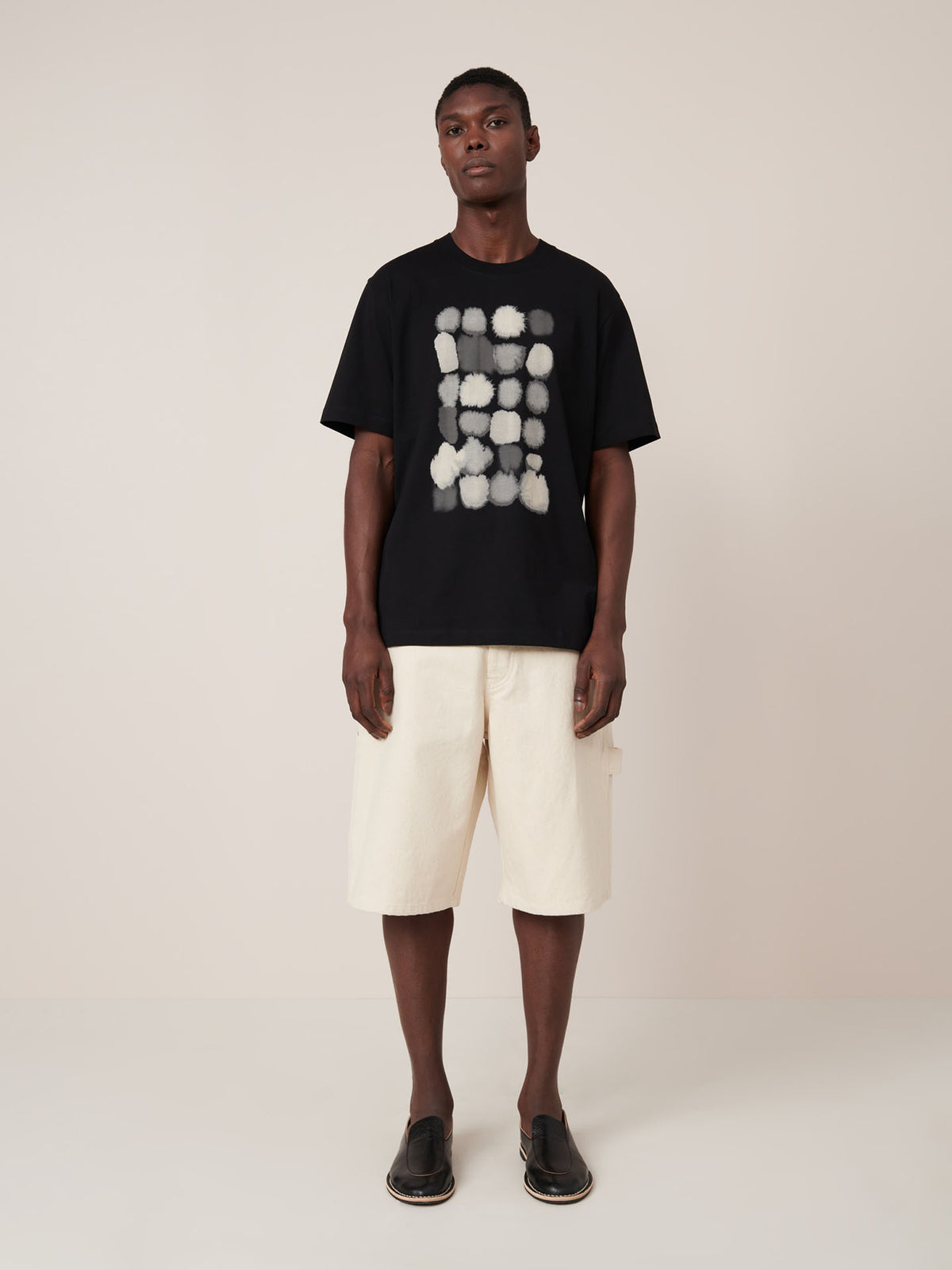 A man stands against a plain background wearing the Kowtow Watercolour Tee in black, featuring a white abstract pattern, paired with cream knee-length shorts and black slip-on shoes.