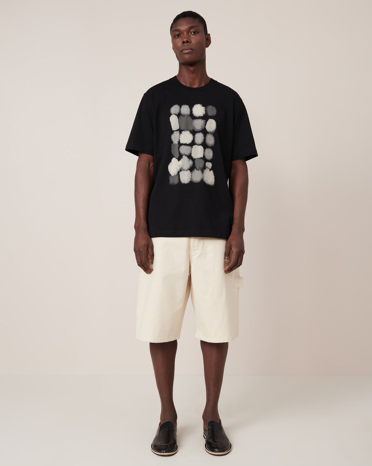 A man wears the Kowtow Watercolour Tee – Black, a standard-fit t-shirt featuring an abstract white and gray rectangular pattern, standing against a plain light background.
