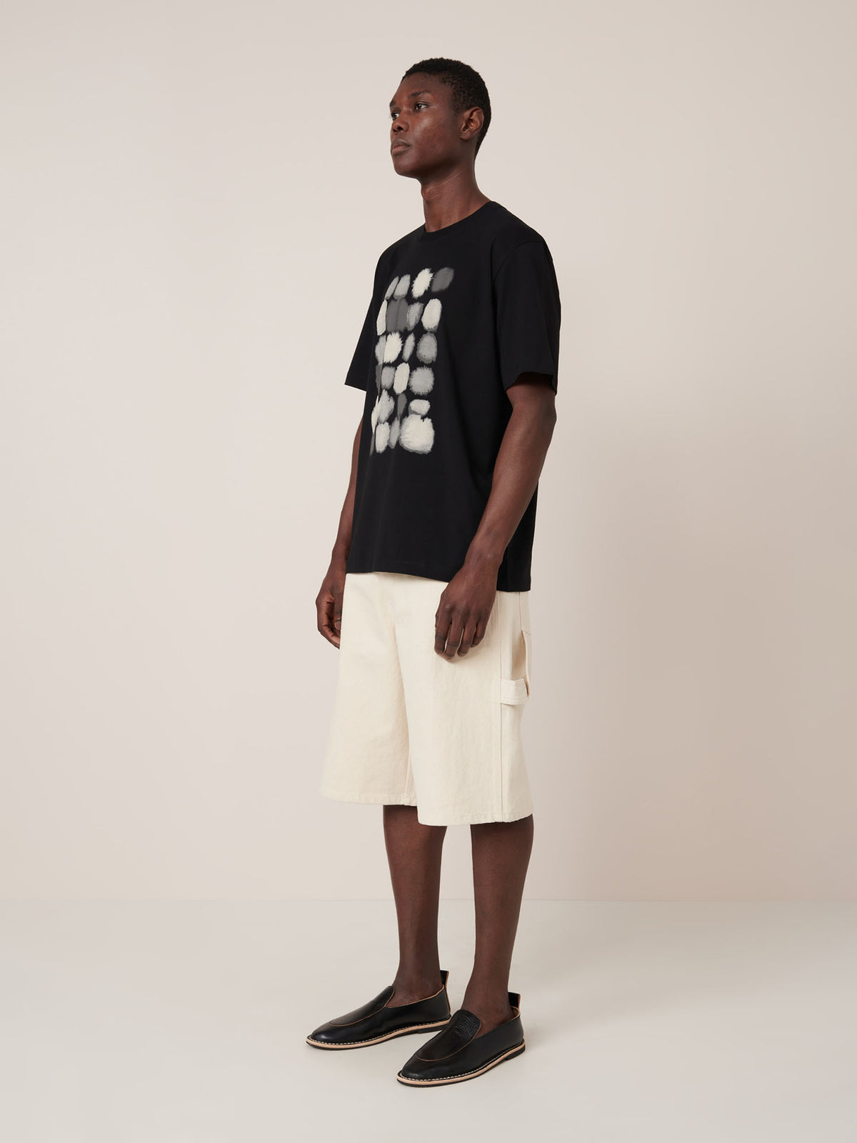 A man stands in a studio wearing the Kowtow Watercolour Tee in black, which features a white abstract pattern and a relaxed fit, styled with cream knee-length shorts and black slip-on shoes.