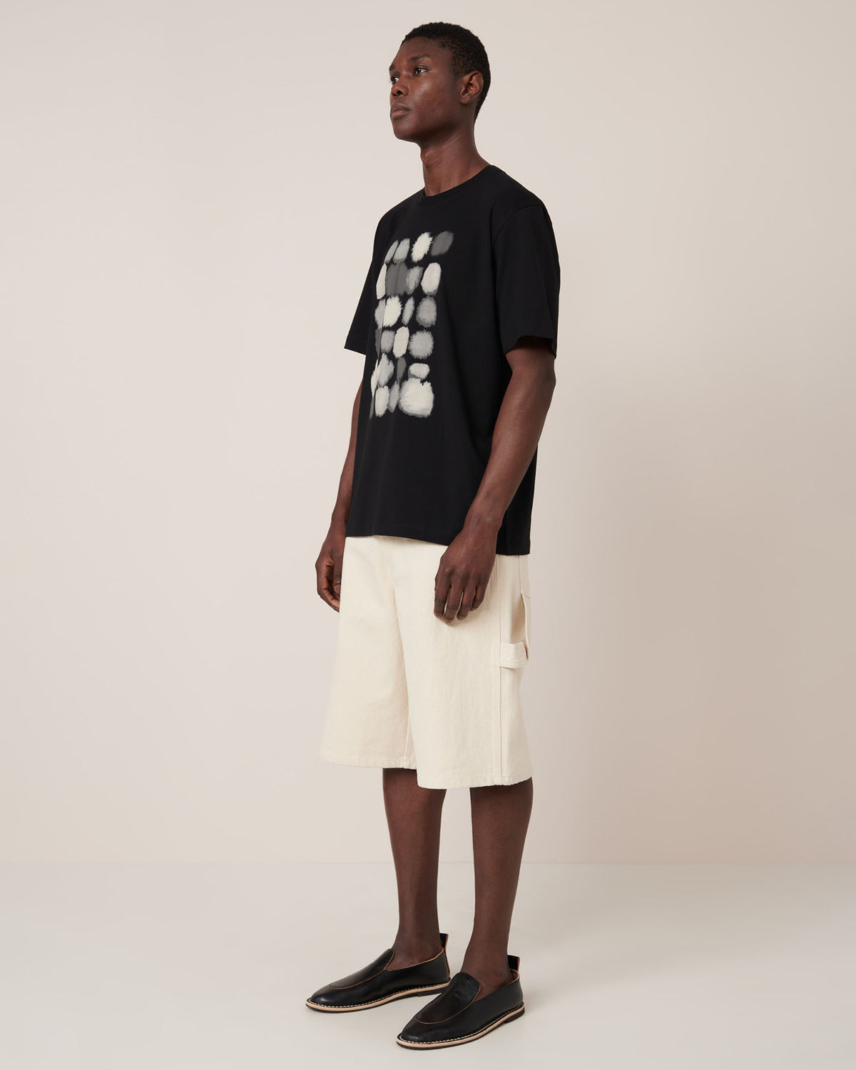 A man stands in a studio wearing the Kowtow Watercolour Tee in black, which features a white abstract pattern and a relaxed fit, styled with cream knee-length shorts and black slip-on shoes.