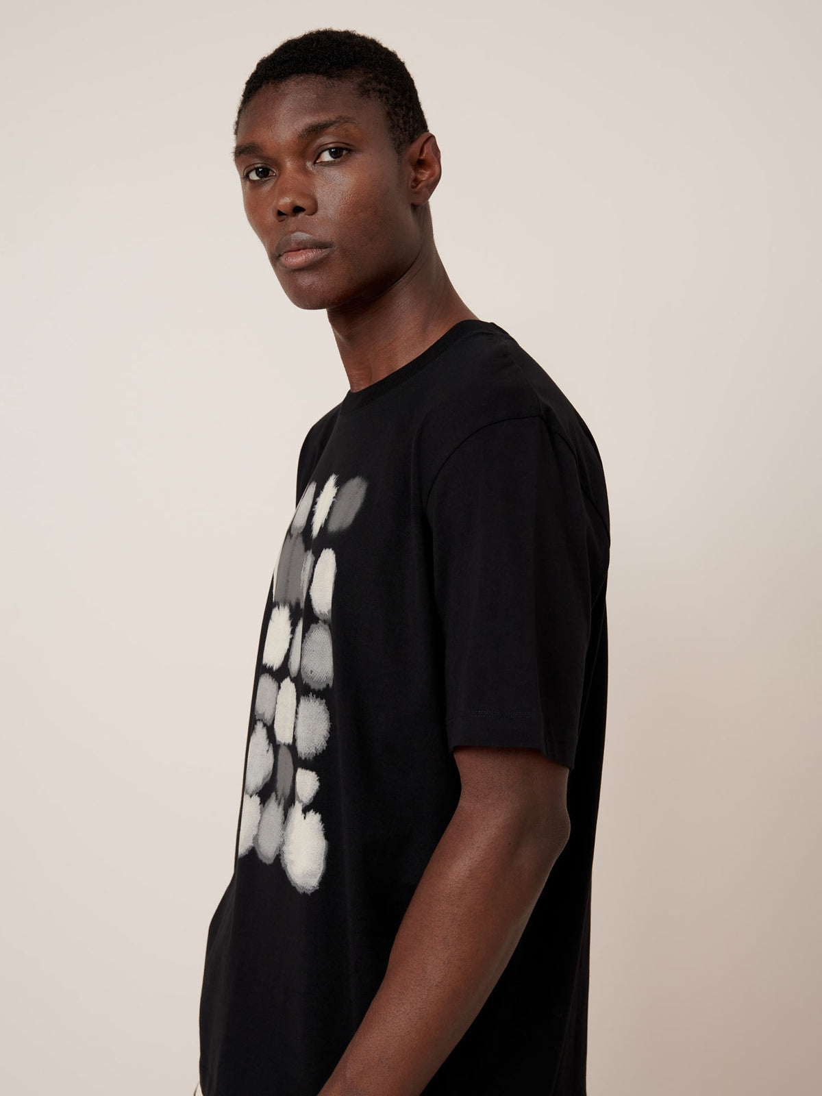 A man stands in profile wearing the Kowtow Watercolour Tee – Black, a relaxed fit T-shirt featuring a white and gray abstract circular design on the front, set against a plain light background.