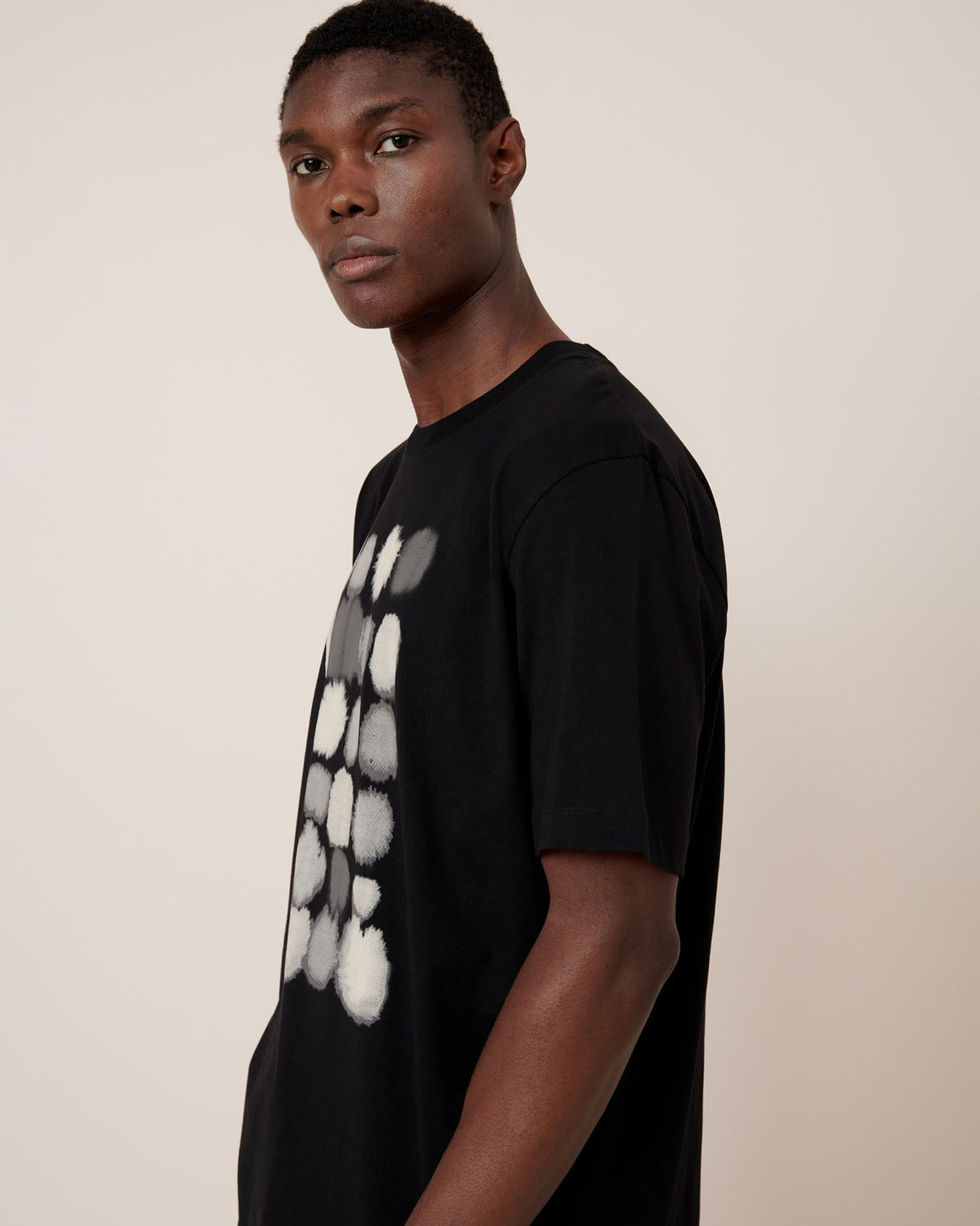 A man stands in profile wearing the Kowtow Watercolour Tee – Black, a relaxed fit T-shirt featuring a white and gray abstract circular design on the front, set against a plain light background.