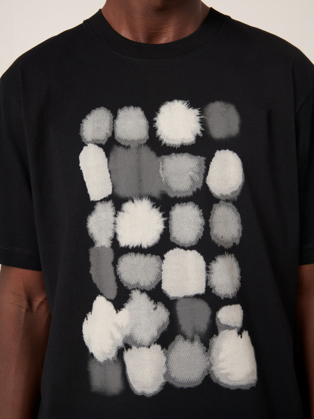 A person wears the Kowtow Watercolour Tee – Black, a relaxed fit t-shirt featuring a grid of irregular white and gray fuzzy shapes on the front.