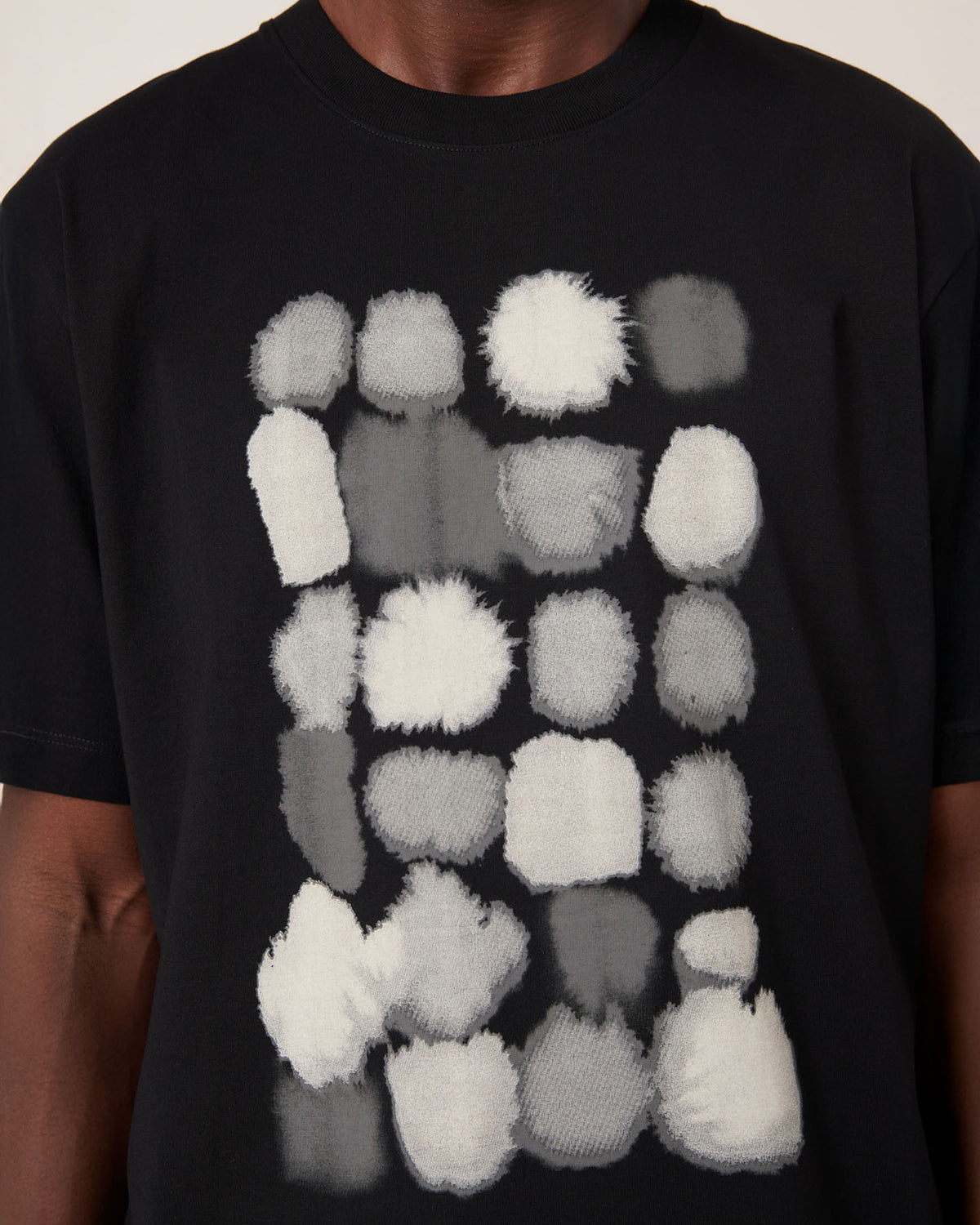 A person wears the Kowtow Watercolour Tee – Black, a relaxed fit t-shirt featuring a grid of irregular white and gray fuzzy shapes on the front.