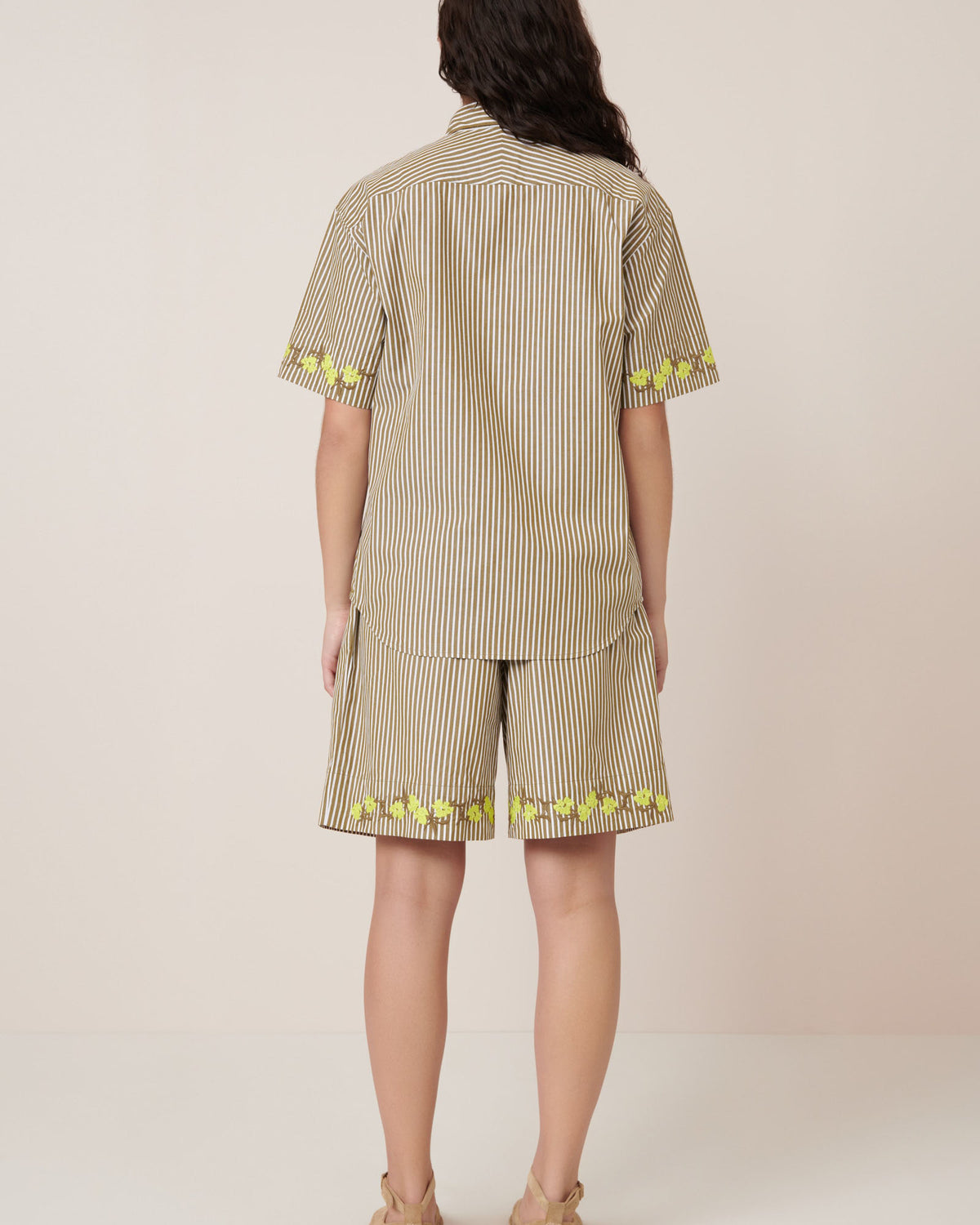 A person faces away, wearing Kowtow's Wildflower Shirt in Treetop Stripe with matching shorts featuring yellow trim, and beige sandals, set against a plain background.