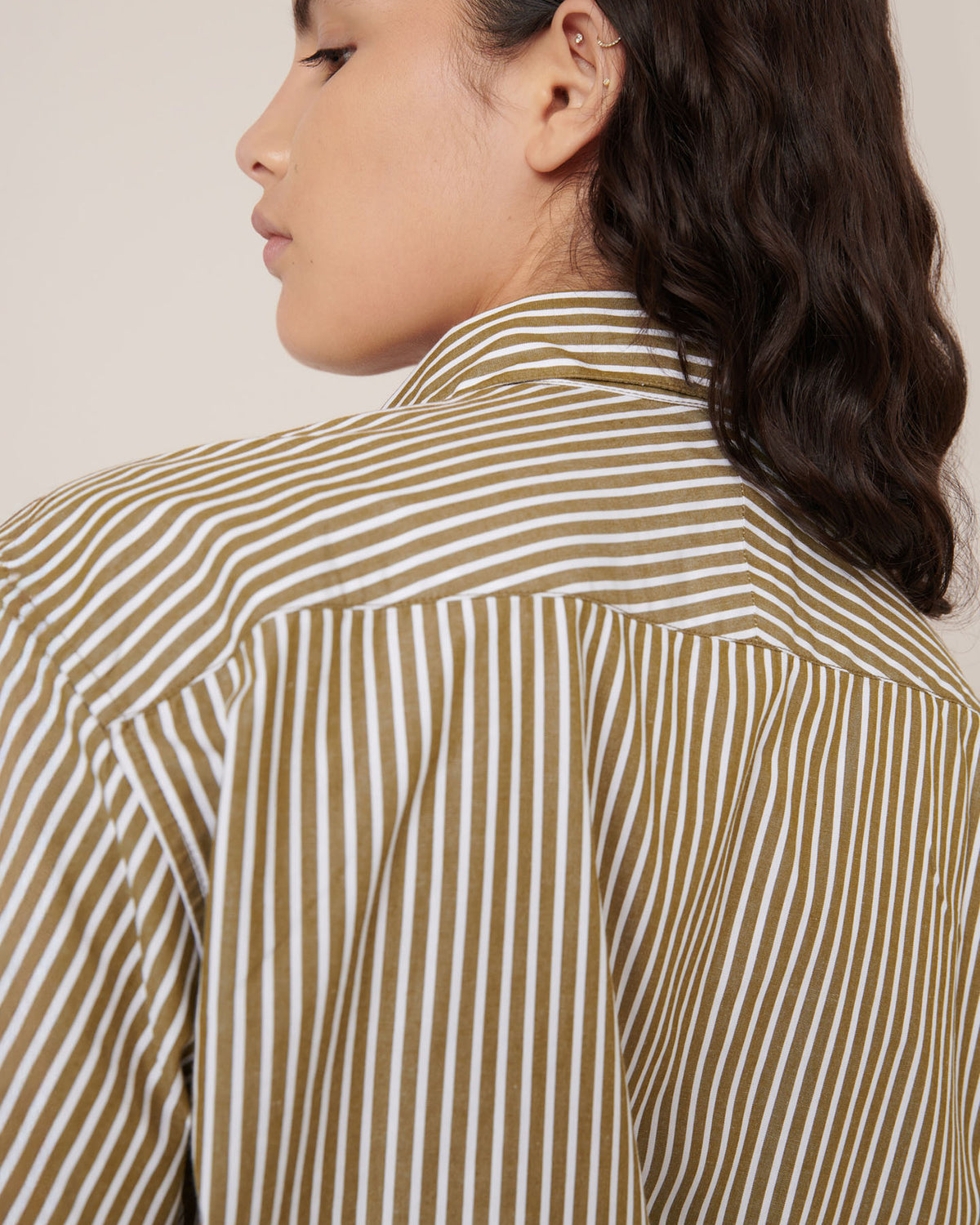 A person with wavy dark hair is shown from the back and side, wearing Kowtow's Wildflower Shirt in Treetop Stripe, featuring a relaxed fit with brown and white vertical stripes.