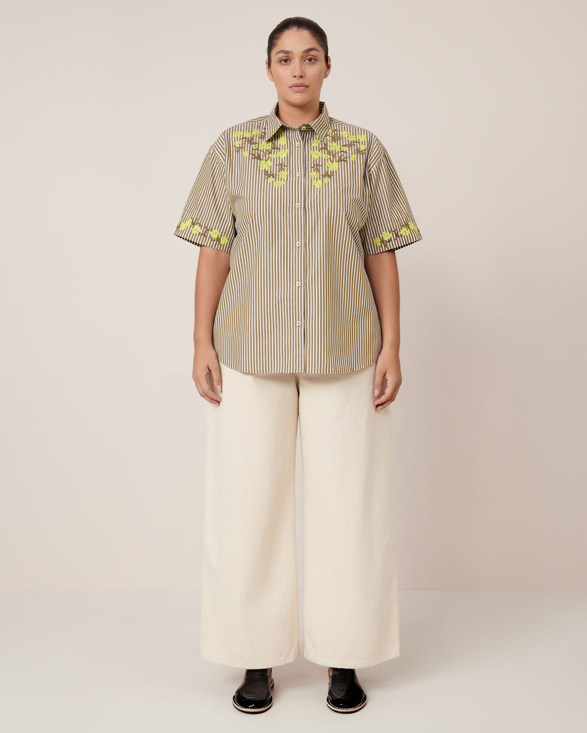 A person stands facing forward in the Kowtow Wildflower Shirt – Treetop Stripe, featuring yellow floral embroidery, paired with relaxed fit wide-leg cream pants and black shoes, against a plain light background.