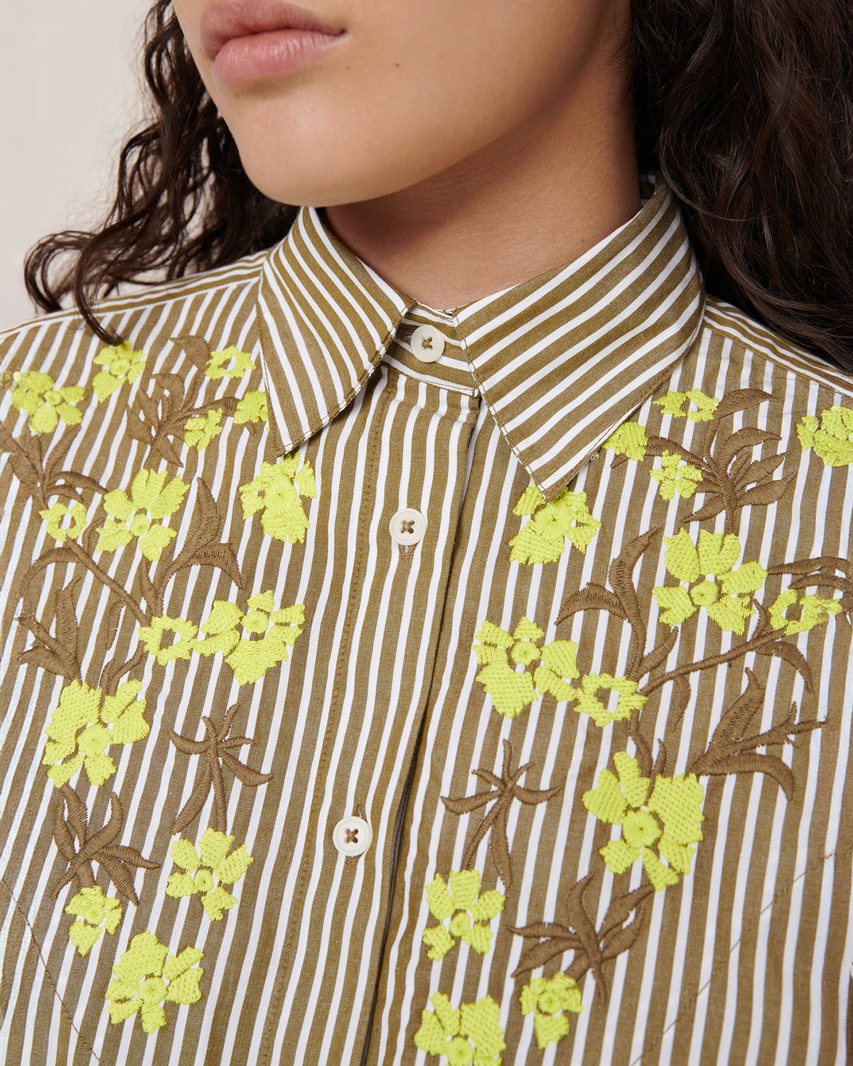 Close-up of a person wearing the Kowtow Wildflower Shirt – Treetop Stripe, a relaxed fit brown and white striped button-up with yellow floral embroidery on the chest and collar.