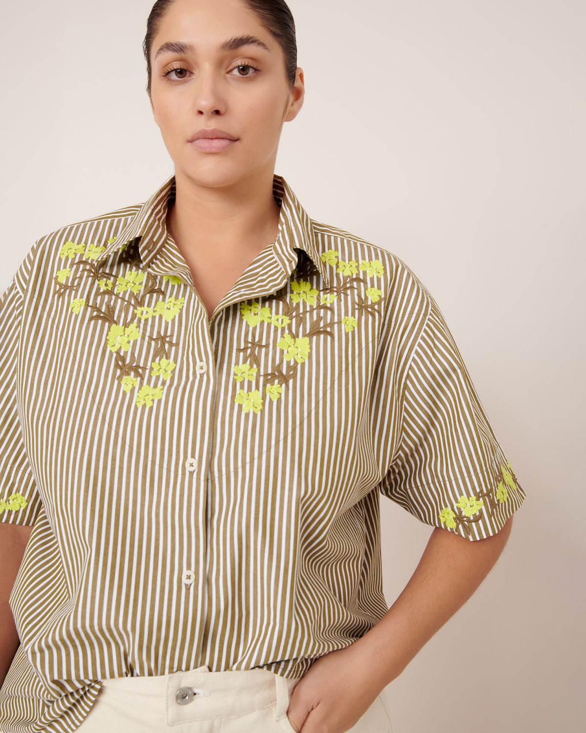 A person stands against a plain background, wearing the Kowtow Wildflower Shirt – Treetop Stripe, a relaxed-fit short-sleeved brown and white striped shirt with yellow floral embroidery, paired with light-colored pants.