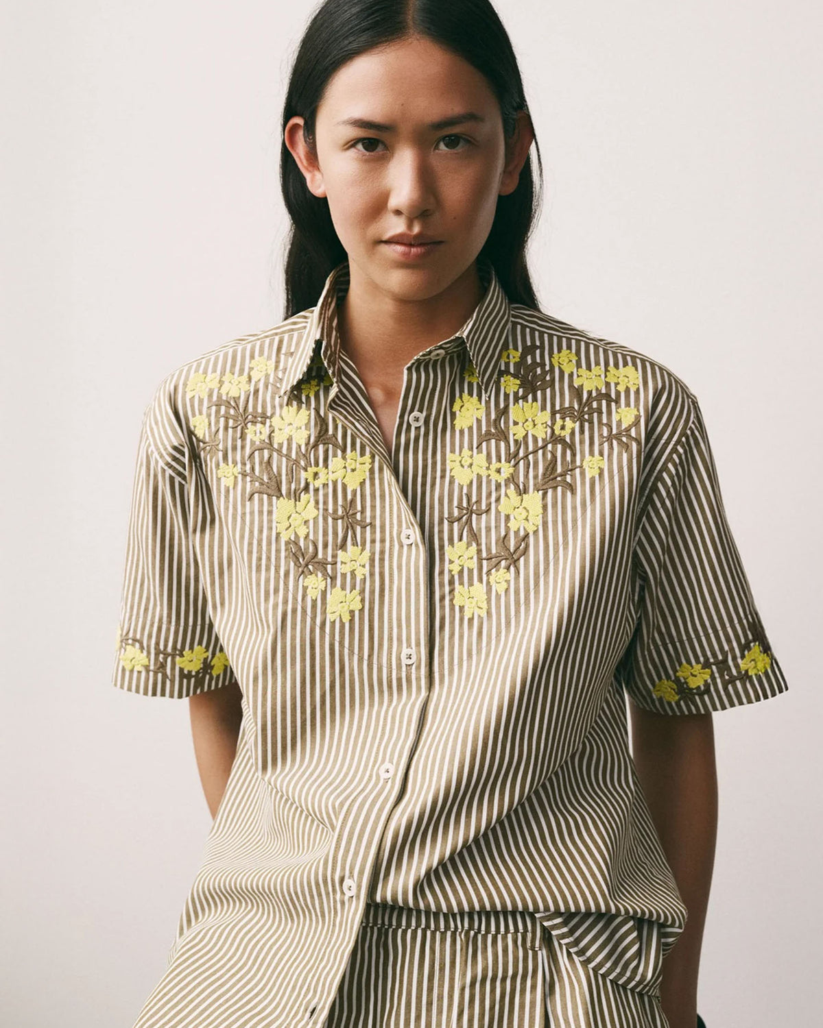 Person with long dark hair wearing the Kowtow Wildflower Shirt – Treetop Stripe, a relaxed fit brown and white striped short-sleeve shirt with yellow floral embroidery, standing against a plain background.
