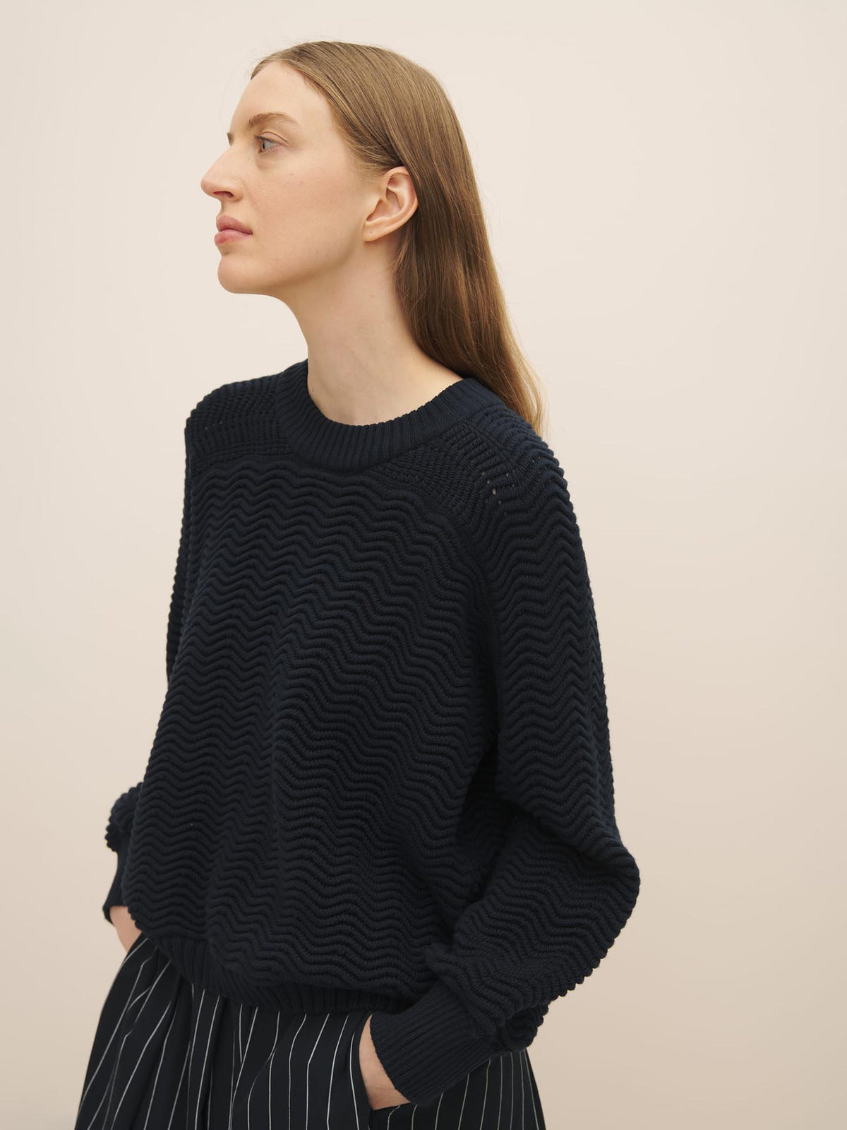 Side profile of a woman with long blonde hair, wearing a Kowtow Zig Zag Crew – Indigo rib knit sweater and striped skirt, against a light beige background.