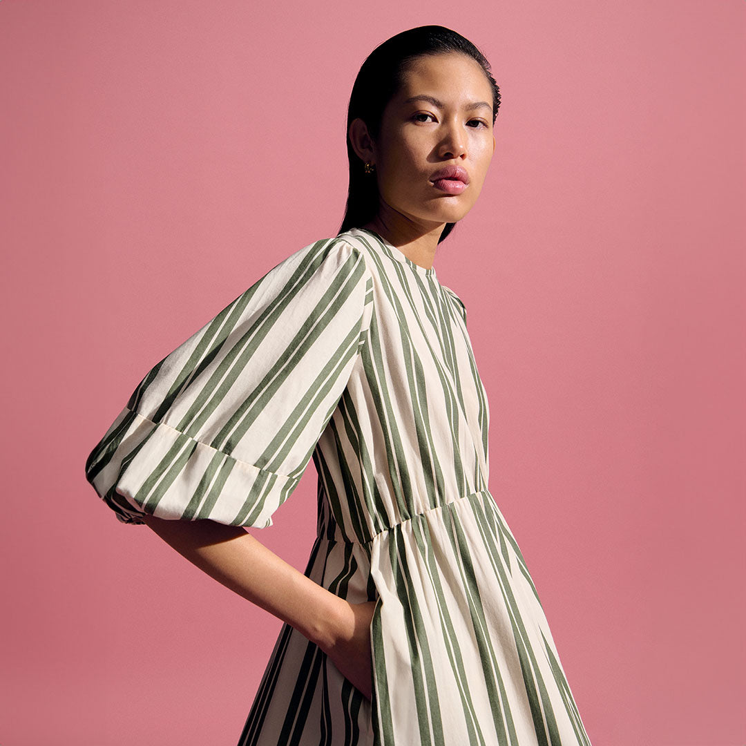 Person in a striped dress with puffed sleeves stands against a pink background, looking towards the camera with hands in pockets.