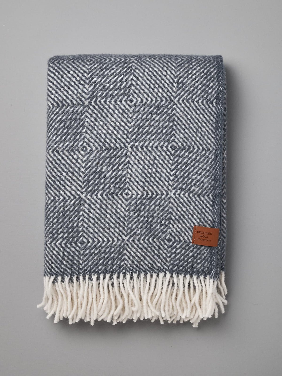 A folded Gooseye Wool Throw – Smokey Blue by Klippan with a geometric pattern and stitched tag is lying on a gray surface. Made from an eco-friendly, sustainable blend, the edges of the throw feature white fringe.