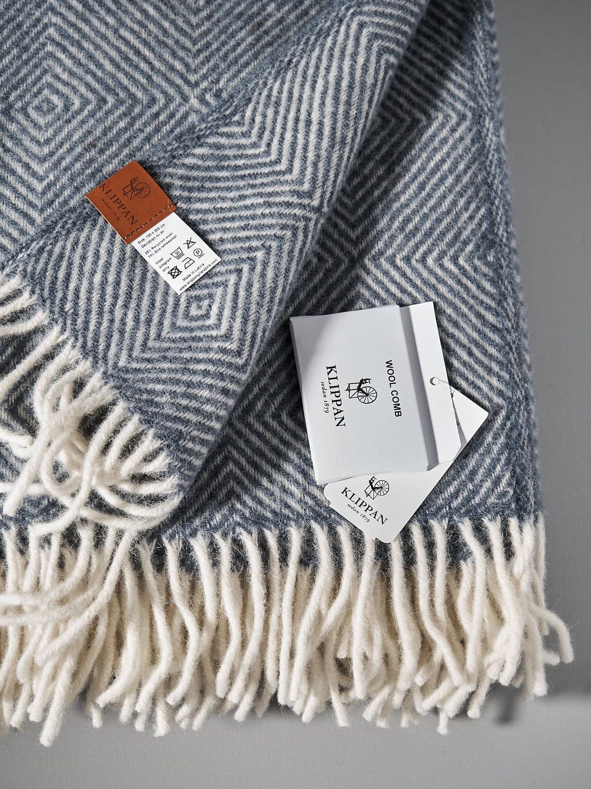 A Gooseye Wool Throw – Smokey Blue made from organic lamb&#39;s wool with a geometric blue and white pattern is shown with its tags. The edges have white fringe. The tags indicate it is from Klippan and provide care instructions.