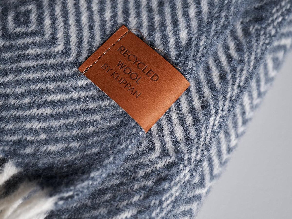 Close-up of a blue and white herringbone-patterned fabric with a brown tag that reads &quot;Gooseye Wool Throw – Smokey Blue by Klippan.&quot; This eco-friendly throw is not only stylish but also sustainable.