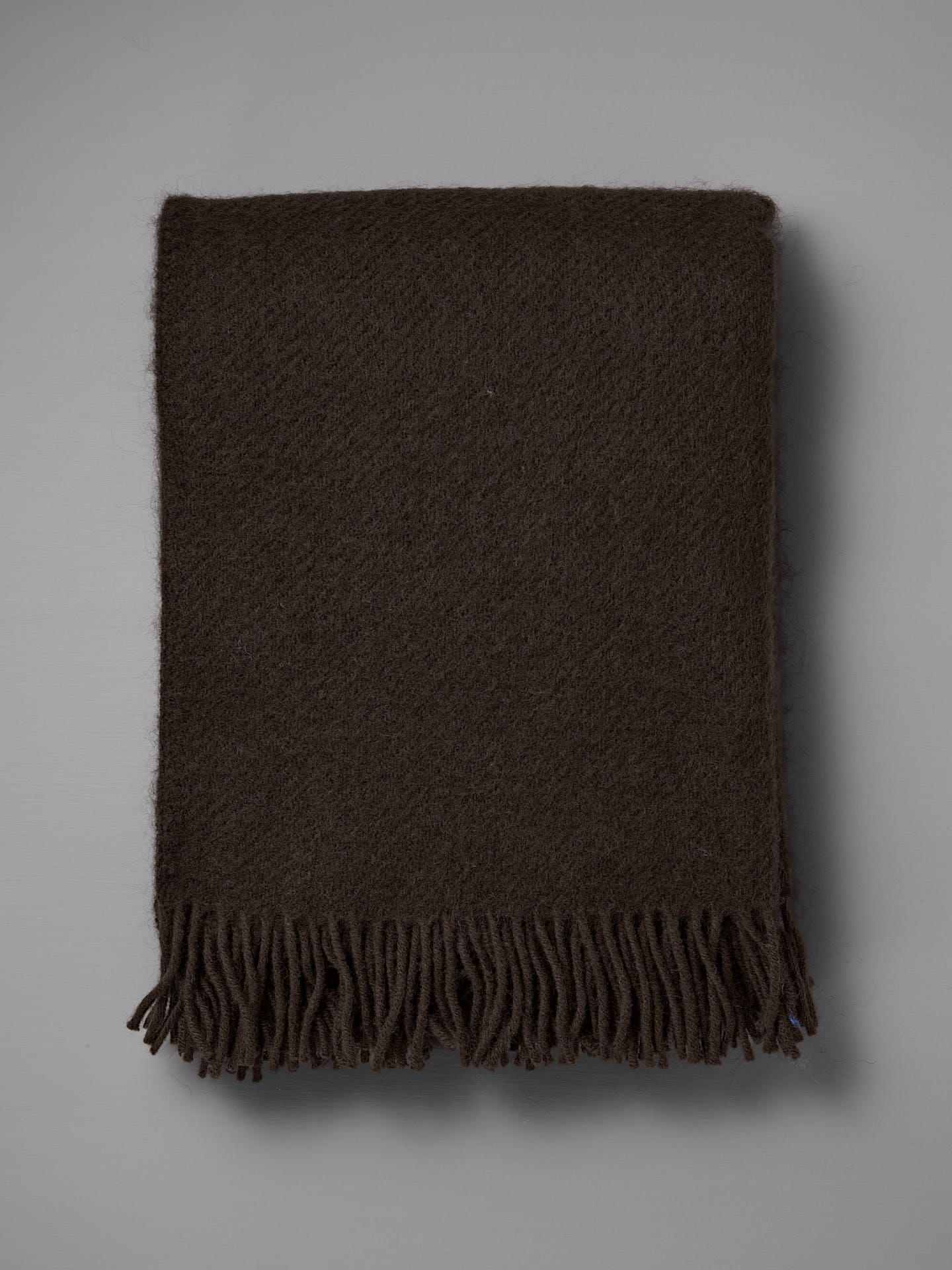 The Gotland Wool Throw – Walnut, featuring hand-knotted tassels and a rich dark brown hue, is neatly folded and displayed on a plain gray background.