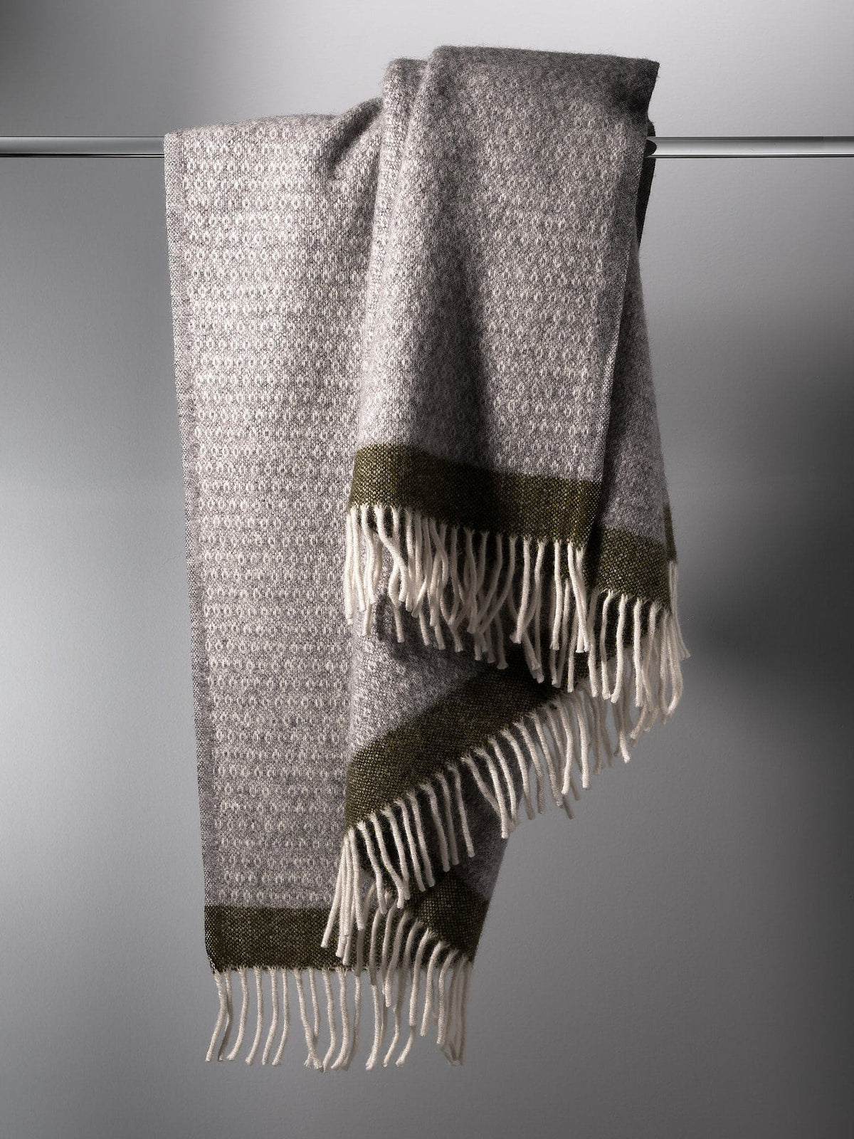 A Hampus Wool Throw – Grey Green with fringes hanging on a hanger. (Klippan)