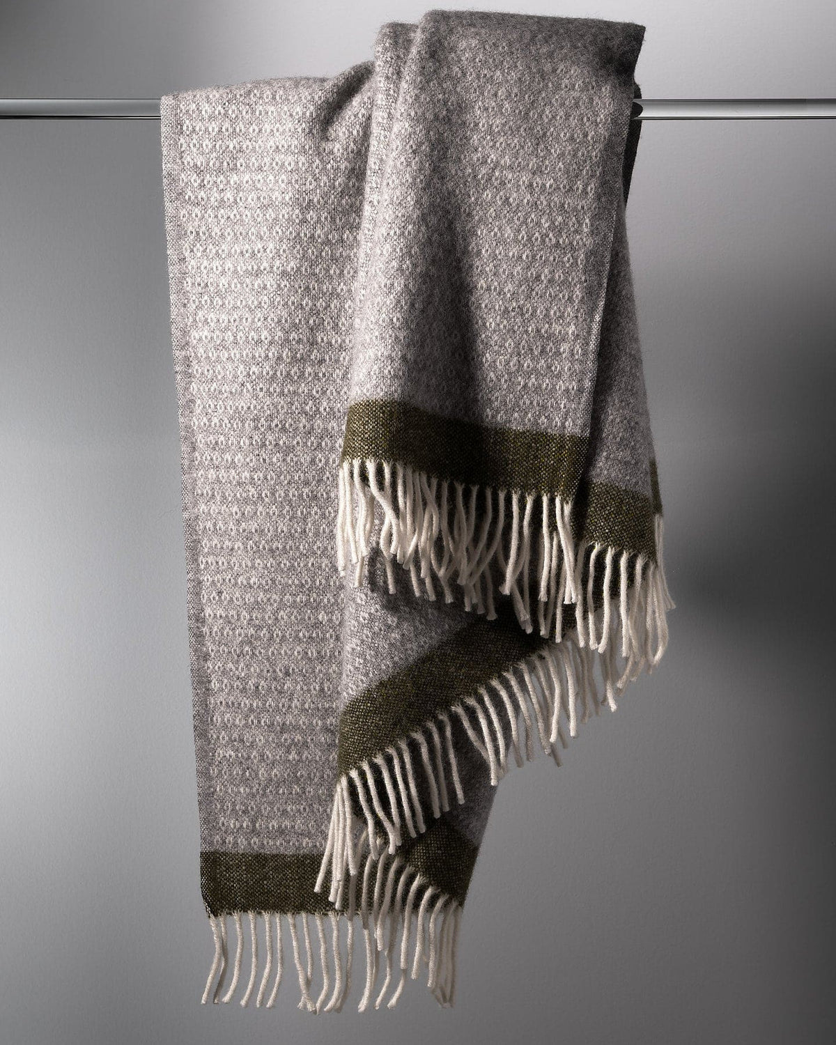 A Hampus Wool Throw – Grey Green with fringes hanging on a hanger. (Klippan)